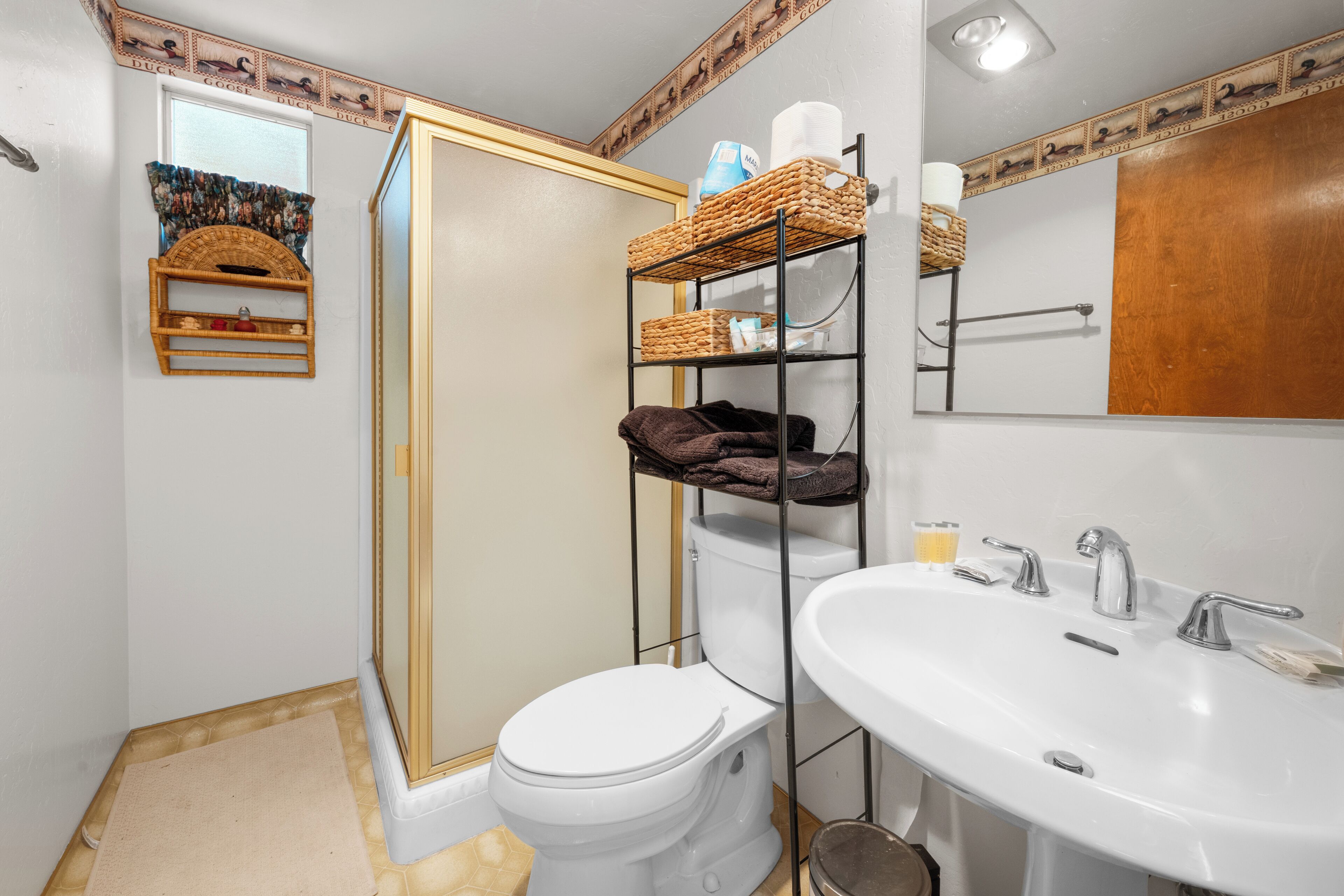 Une salle de bain avec des toilettes blanches, un lavabo blanc et une douche dotée d’une porte en verre. On y trouve des paniers en osier sur une étagère en métal au-dessus des toilettes, ainsi qu’un porte-serviettes sur lequel est accrochée une serviette.