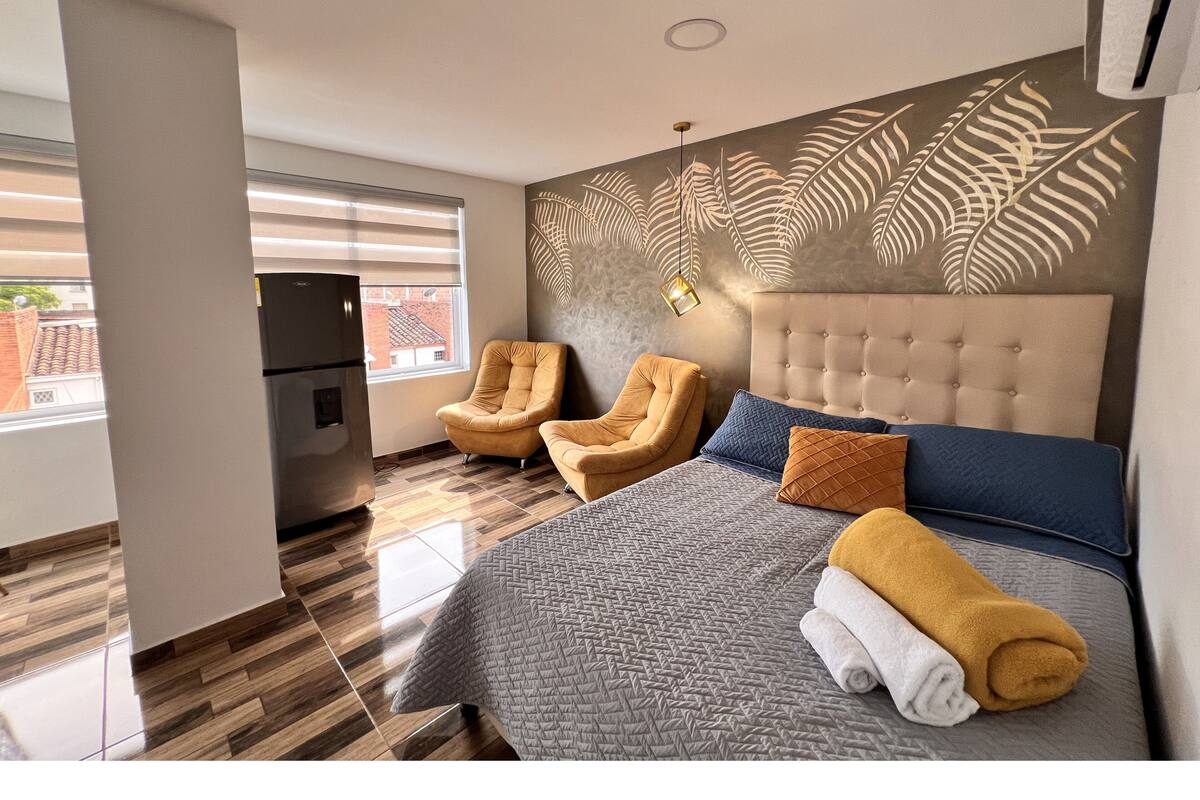 Une chambre d’hôtel moderne équipée d’un lit, de deux fauteuils, d’un réfrigérateur et d’une grande fenêtre.