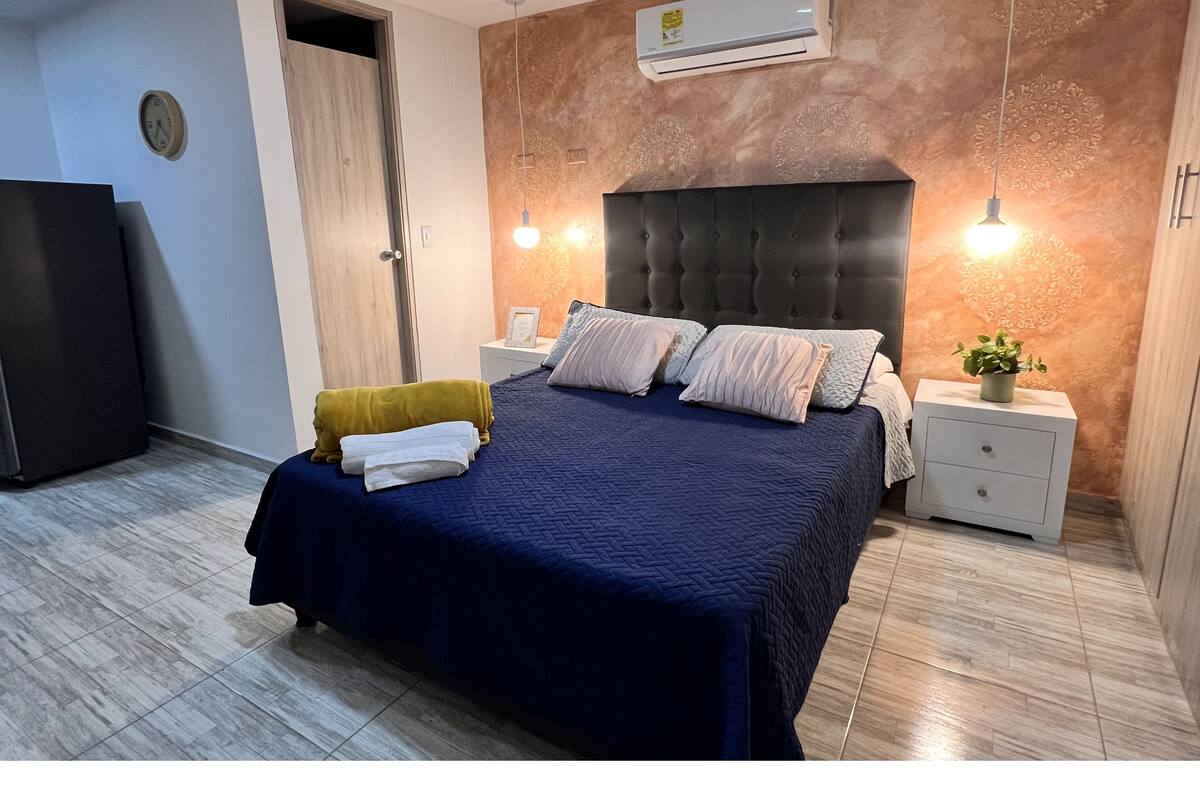 Une chambre à coucher comprenant un lit, des tables de chevet, une commode, un climatiseur fixé au mur, une horloge et une porte.