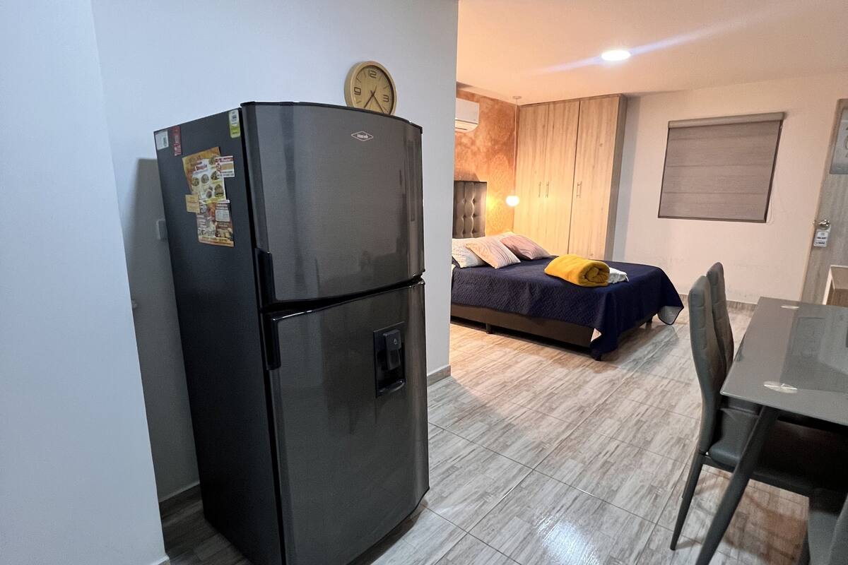 Une cuisine compacte équipée d’un réfrigérateur, un lit avec une couverture bleue et un coin repas comprenant une table et des chaises.