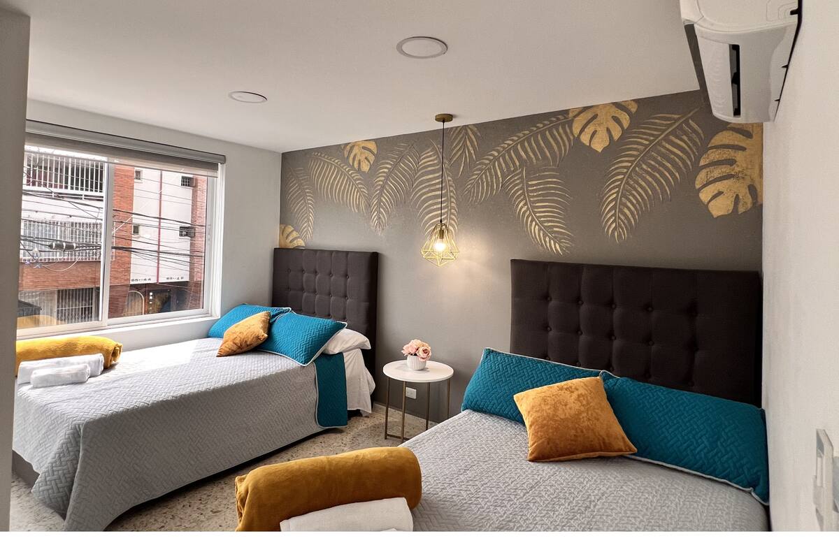 Une chambre d’hôtel avec deux lits, une petite table ronde, un climatiseur fixé au mur et une fenêtre donnant sur des immeubles.