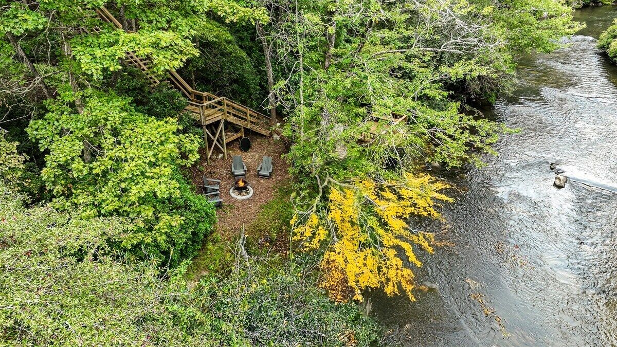 Ett avskilt utomhusområde med sittplatser och en eldstad, omgivet av tät skog och en flod.