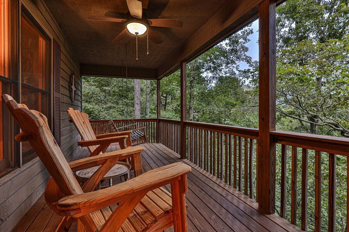 A wooden deck with a ceiling fan, wooden chairs, and a view of trees.