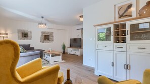 Ein Wohnzimmer mit einer Couch, einem gelben Sessel, einem Fernseher, einer Vitrine mit Weingläsern und einer Steinwand.