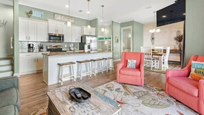 A modern kitchen with white cabinets, stainless steel appliances, and a central island with bar stools. There is a dining area with a table and chairs, and a living room with a sofa and a coffee table. - Little Lagoon Cottage for 8 - Free Activities (Gulf Shores)