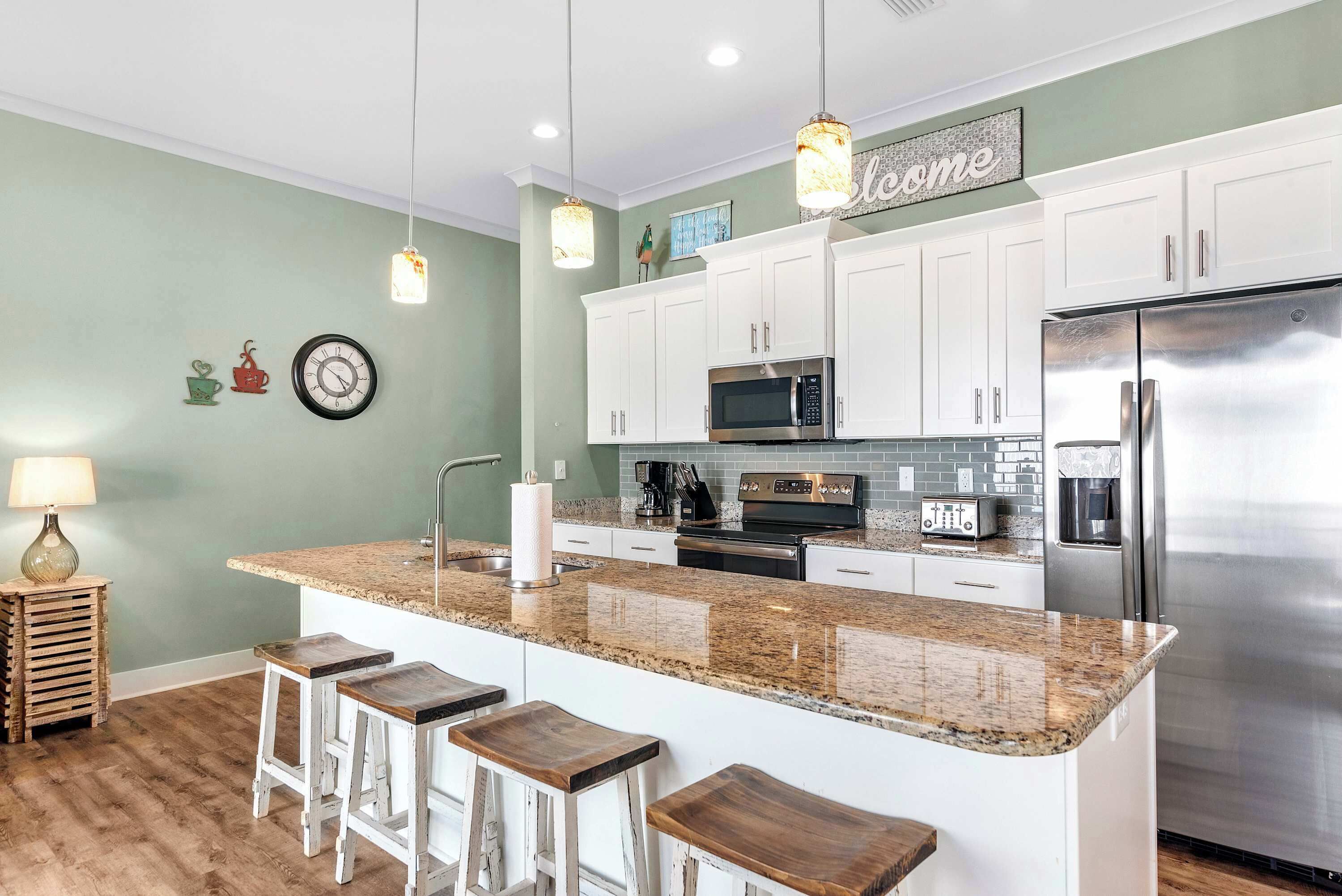 A modern kitchen with white cabinets, a stainless steel refrigerator, a granite countertop, and wooden bar stools.