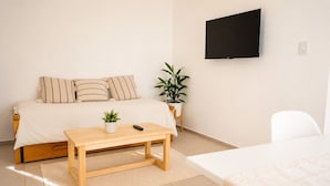 Un dormitorio con cama, mesita de noche con una planta, televisor montado en la pared y una silla.