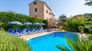 A pool with lounge chairs and umbrellas, surrounded by greenery and a building in the background.