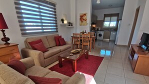 Ein Wohnzimmer mit einer Couch, einem Couchtisch und einem Essbereich. Im Hintergrund ist eine Küche mit Geräten und einem Fenster zu sehen.