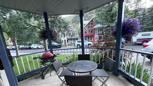 Une terrasse couverte avec une table ronde et des chaises, un barbecue et une vue sur une rue résidentielle.