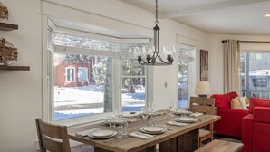 A dining area with a wooden table set for six, large windows with a view of a snowy landscape, and a red sofa in the background. - Smuggler Mine — Modern Pet-Friendly Retreat Near Slopes (Telluride)