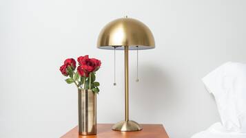 Una mesita de noche de madera con una lámpara de bronce, un florero con flores rojas y una cama blanca.