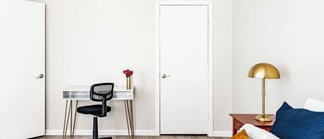 A bedroom with a bed, a desk with a chair, a door, and a nightstand with a lamp.