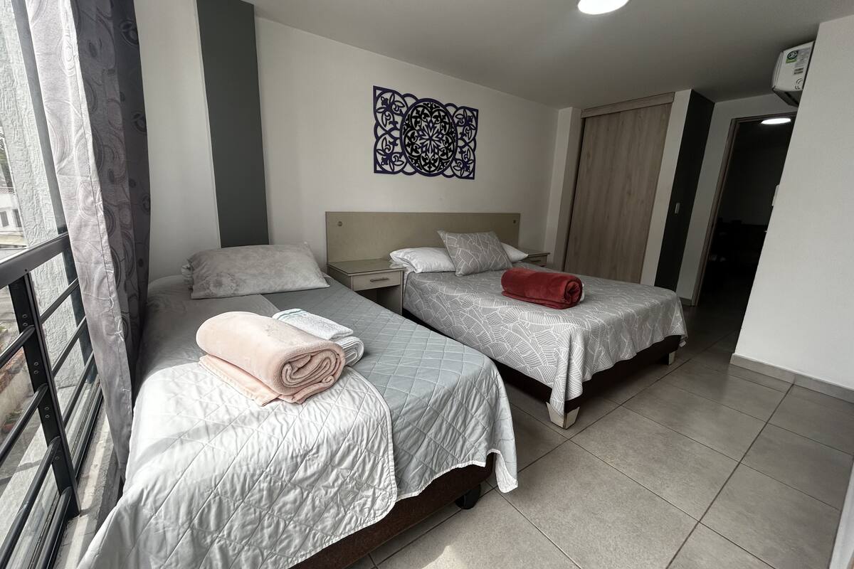 A hotel room with two beds, a window with curtains, a decorative wall piece, and a ceiling light.