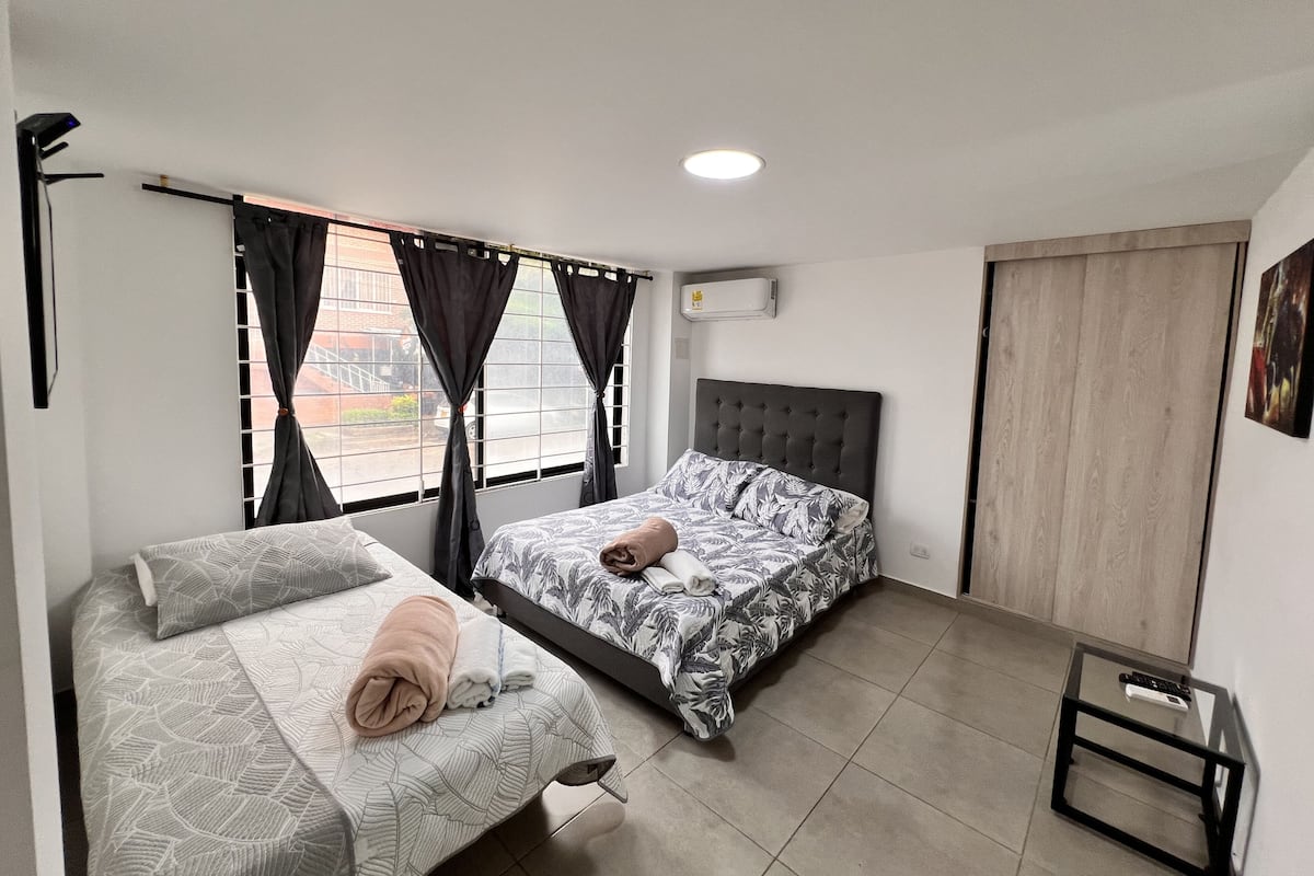 A bedroom with two beds, a window with curtains, an air conditioner, and a small table.