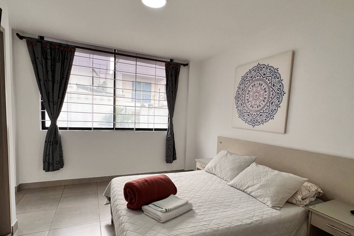 A bedroom with a bed, curtains, a bedside table, a nightstand, and a wall art piece.
