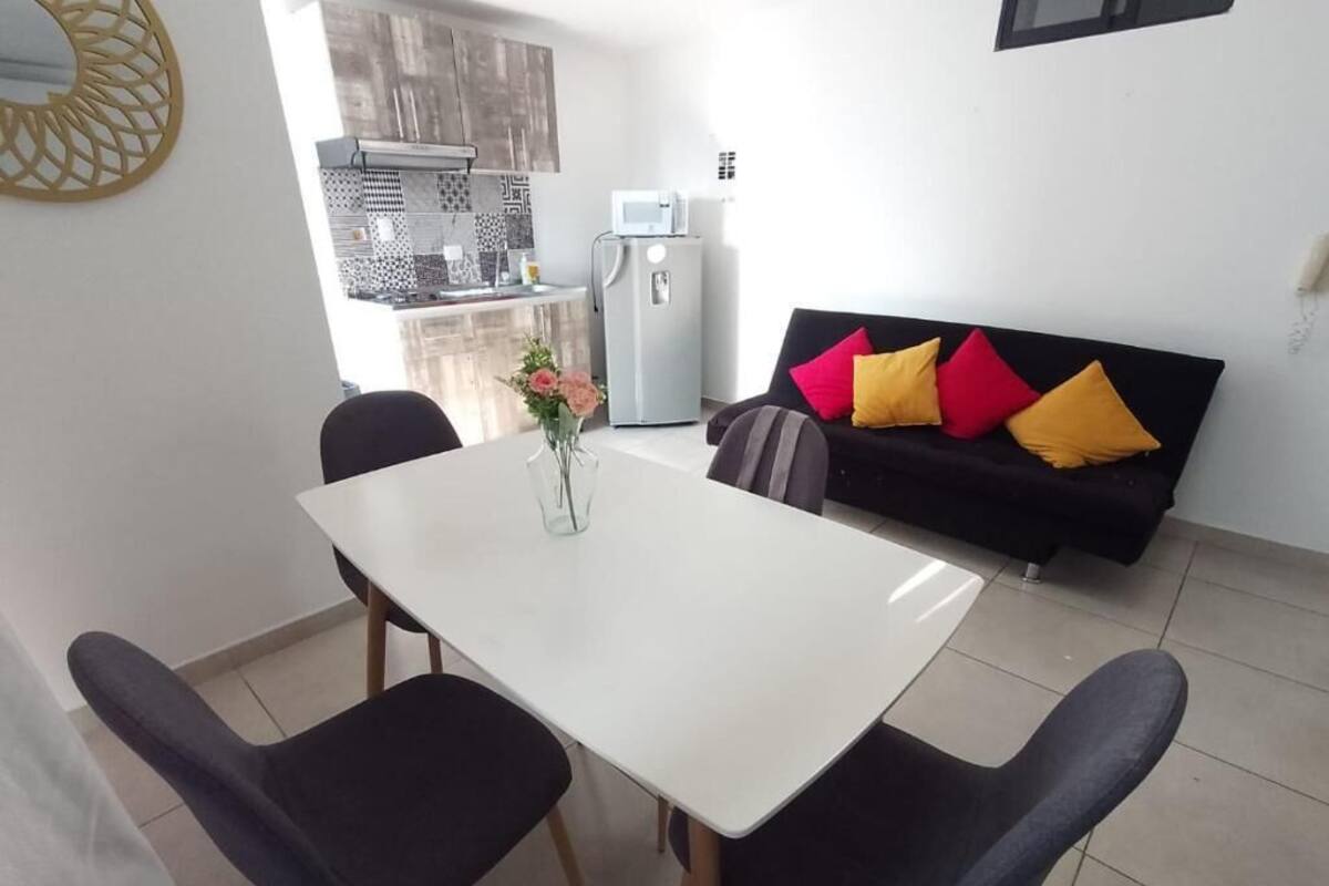 A dining area with a white table and four chairs, a black sofa with colorful cushions, and a kitchen in the background.