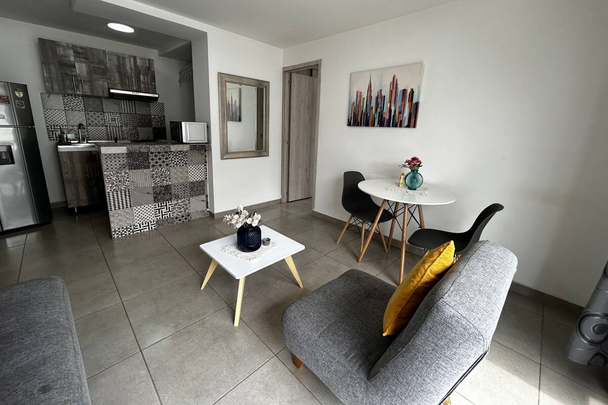 A modern living space with a kitchen, dining area, and a gray sofa with a yellow pillow.
