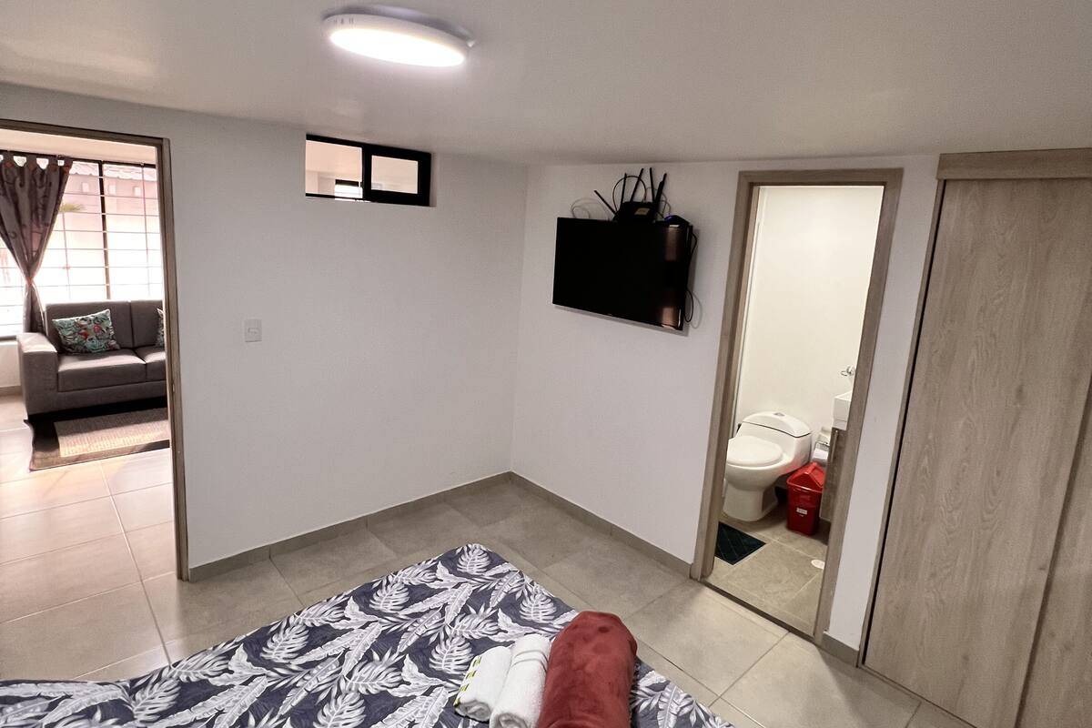 A bedroom with a bed, a TV mounted on the wall, a door leading to a bathroom, and a window with curtains.