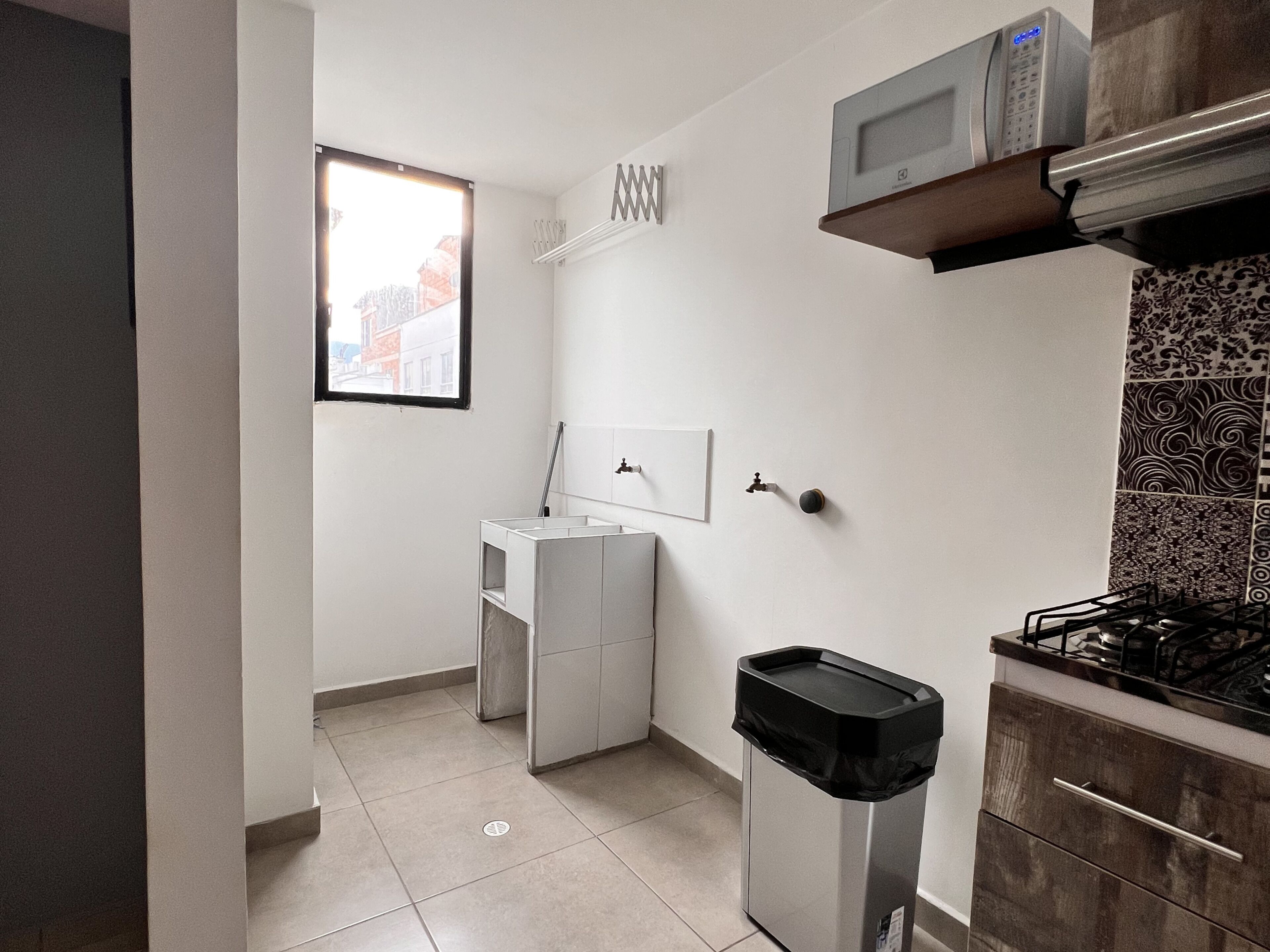 A compact kitchen with a microwave, a gas stove, a trash bin, and a window.