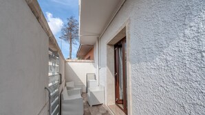 Un estrecho pasillo exterior con mobiliario de mimbre blanco, baranda metálica y una puerta con panel de vidrio.