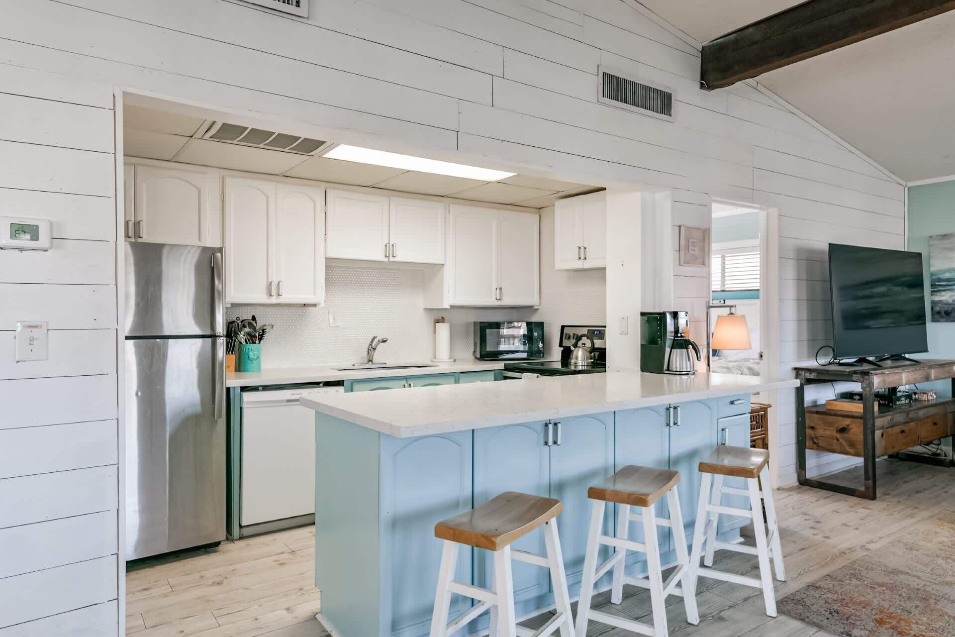 A modern kitchen with white cabinets, a stainless steel refrigerator, a central island with stools, and a TV in the adjacent living area.