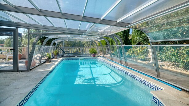 Una piscina con cerramiento de vidrio, rodeada de plantas en macetas y vistas a zonas verdes.