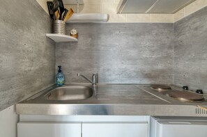 Una cocina con un lavamanos de acero inoxidable, dos hornallas y un estante blanco con utensilios de cocina.