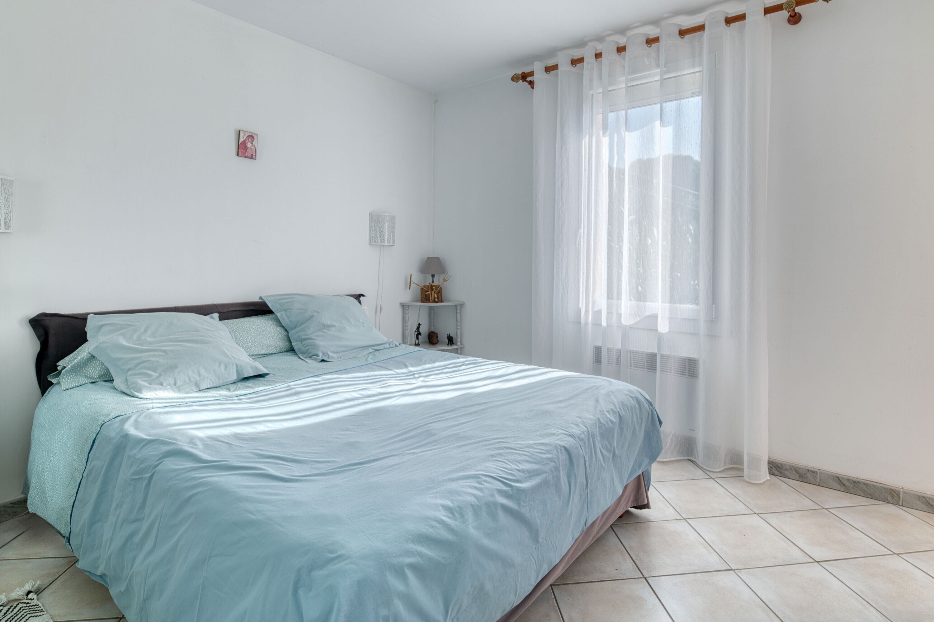 Ein Schlafzimmer mit einem Bett, einem Nachttisch, einer Lampe und einem Fenster mit transparenten Vorhängen.