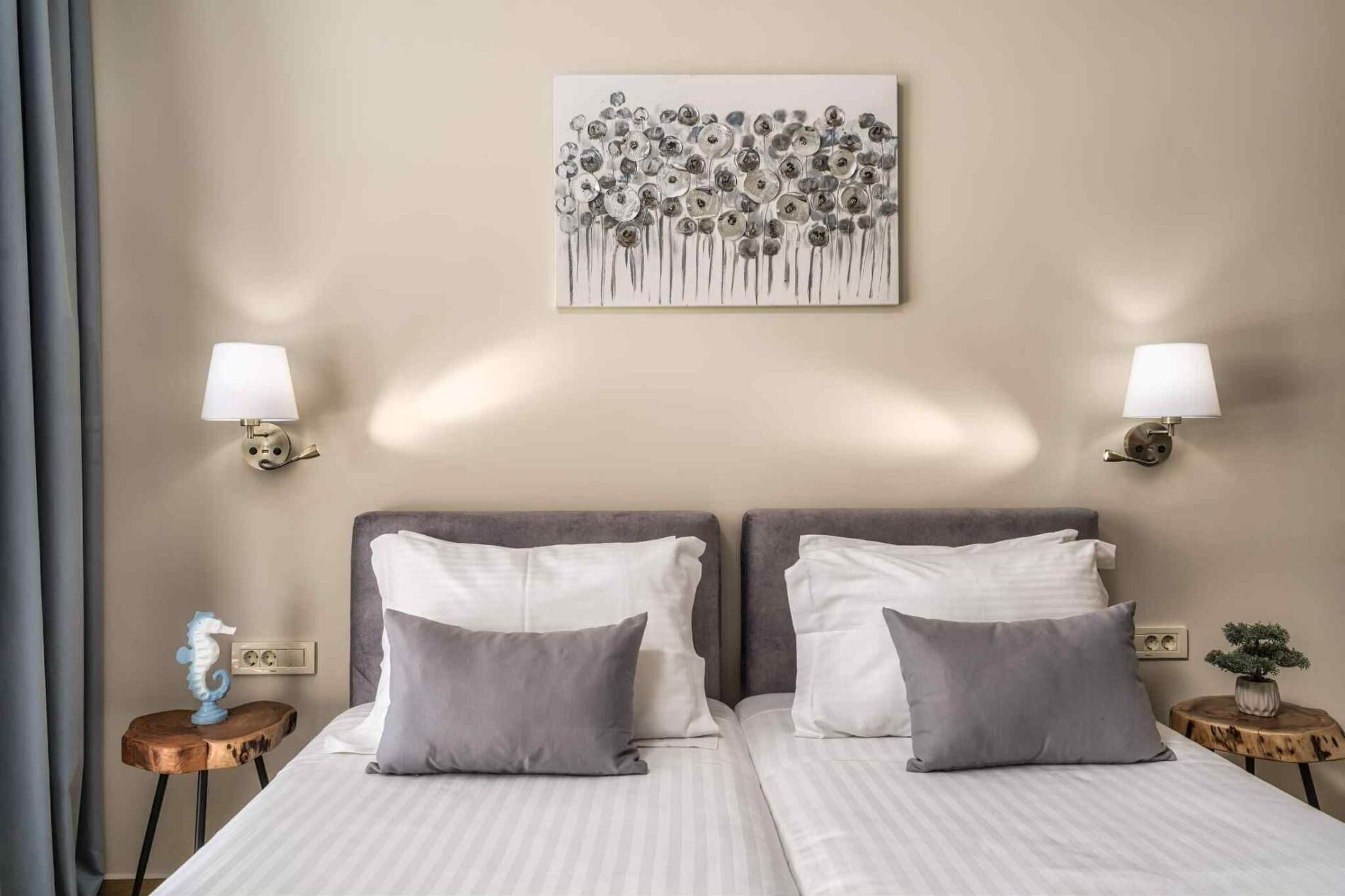 Een tweepersoonsbed met twee kussens, een aan de muur bevestigd kunstwerk, twee wandlampen, een klein houten tafeltje met een decoratief object en een potplant.