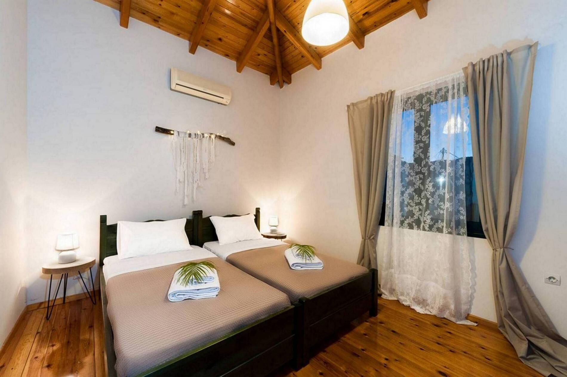 Een slaapkamer met twee bedden, houten vloeren, een plafondventilator, een airconditioning en een raam met gordijnen, plus een wandlamp.