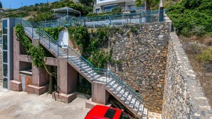 Un bâtiment moderne doté d’un mur de soutènement en pierre, d’un escalier avec des rampes en métal et d’une voiture rouge garée devant.