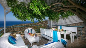 Un espace extérieur abrité, aménagé avec des meubles en osier, une table dressée avec des assiettes et des tasses, et une vue sur la mer.