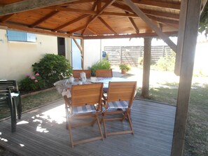 Eine überdachte Terrasse mit einem Holztisch und Stühlen, umgeben von Topfpflanzen und Blick auf einen Zaun.