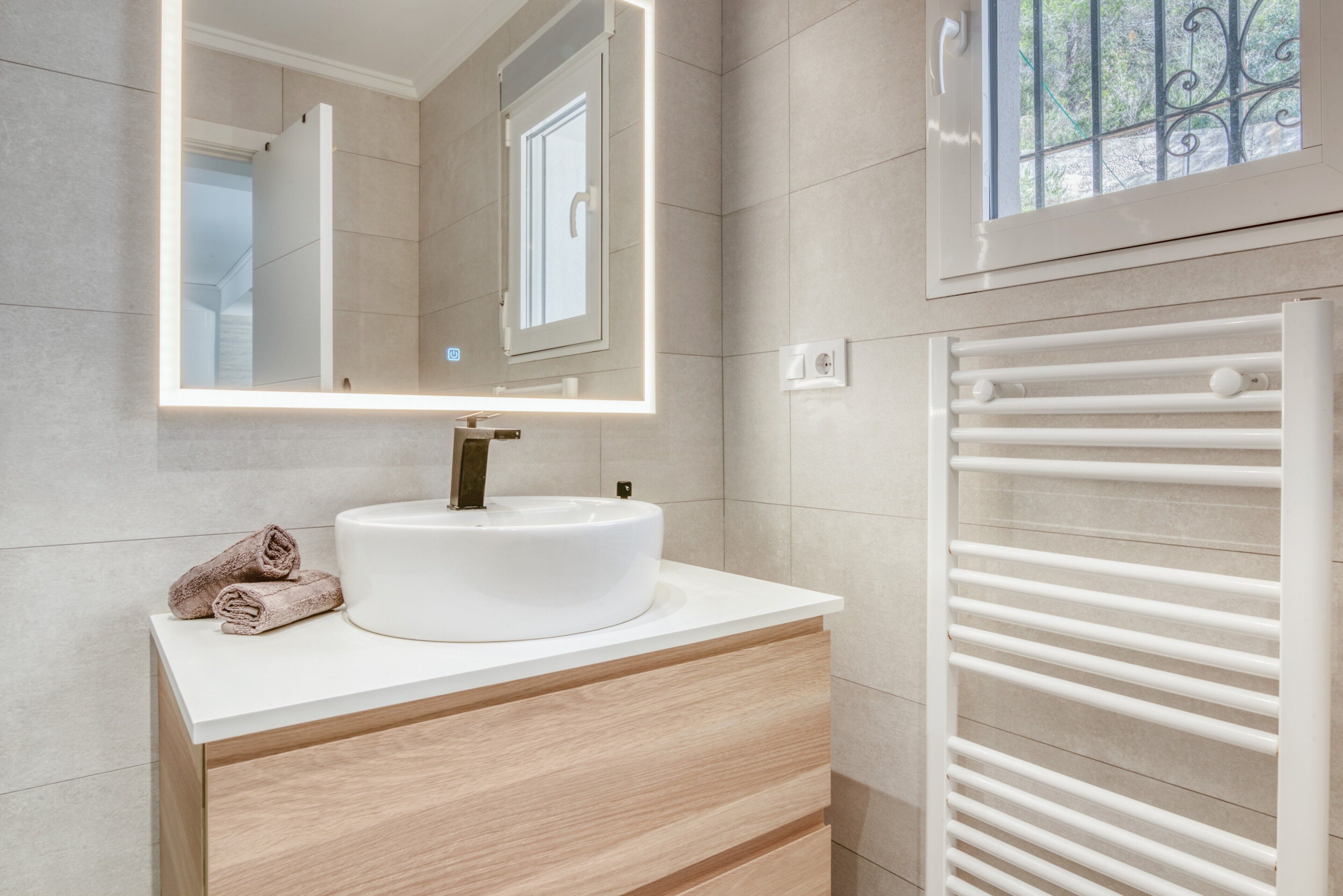 Une salle de bain moderne avec un lavabo rond, un miroir doté d’un éclairage intégré, un porte-serviettes et une fenêtre offrant une vue agréable.