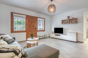 Ein modernes Wohnzimmer mit einer Couch, einem kleinen Tisch mit einer Pflanze, einem Fernseher auf einem weißen Schrank und großen Fenstern.