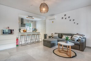 Ein modernes Wohnzimmer mit einem grauen Sofa, einem kleinen runden Tisch und einer Küchenzeile mit Mikrowelle und Hockern.