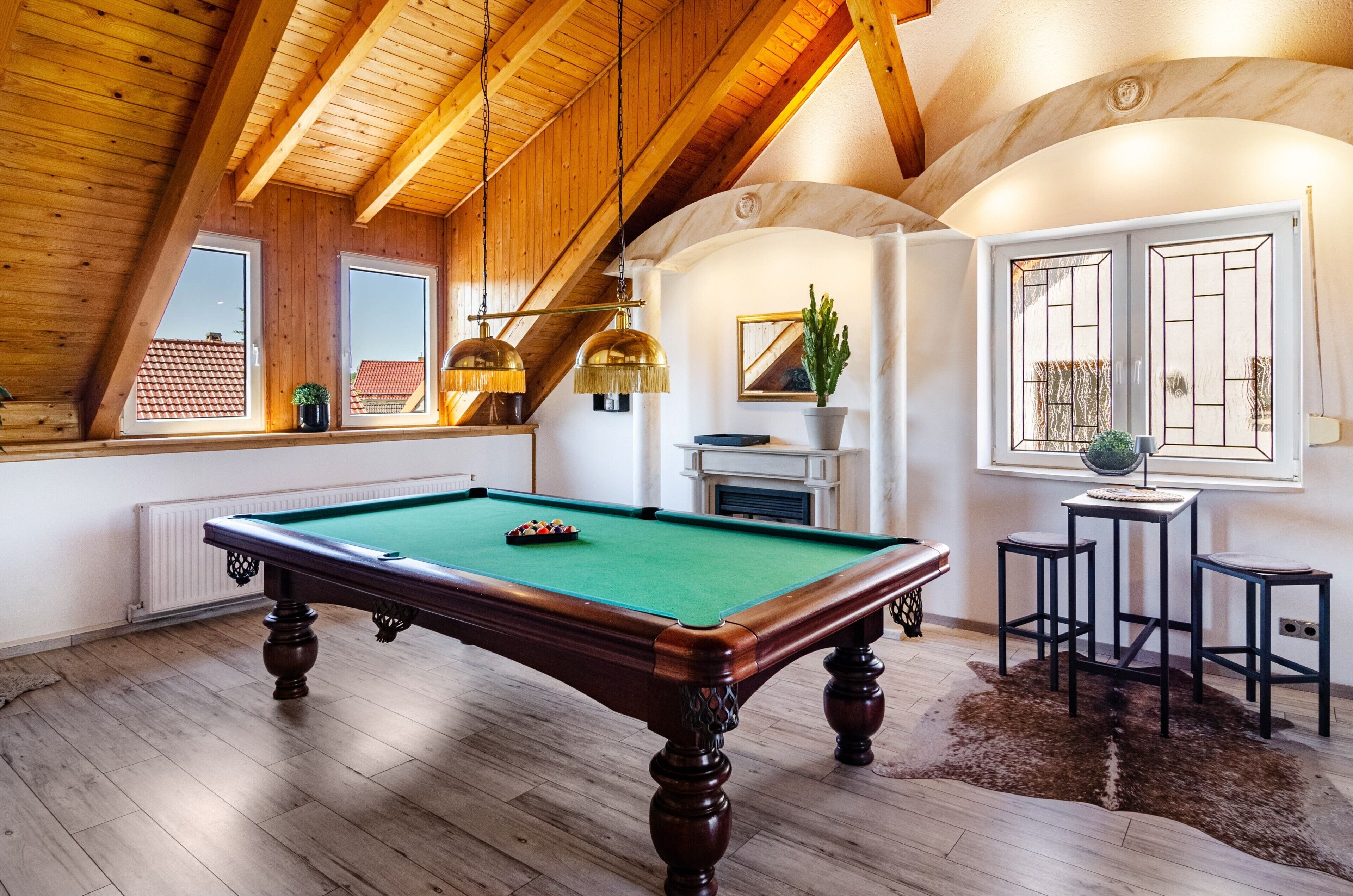 Une table de billard dans une pièce avec des poutres en bois, une cheminée et une vue sur un toit recouvert de tuiles rouges.
