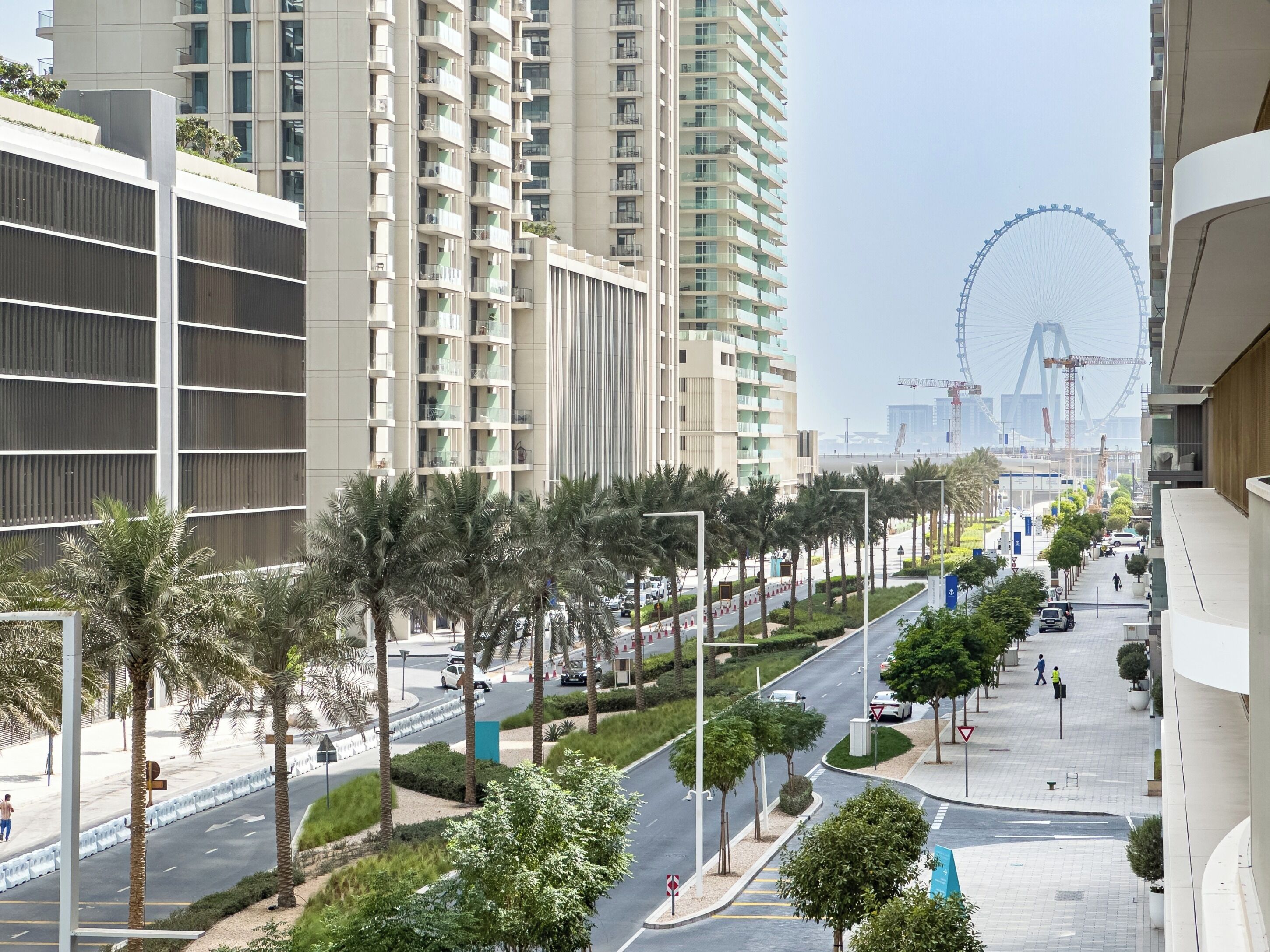 Une rue moderne en milieu urbain, bordée d’immeubles résidentiels imposants, de palmiers et avec une grande roue en arrière-plan.