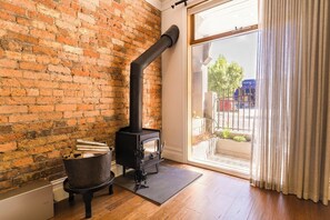 A room with a brick wall, a wood-burning stove, a window with curtains, and a door leading outside.