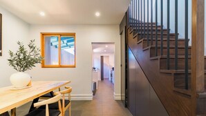 Ein modernes Interieur mit einem Holzesstisch, Stühlen, einer Topfpflanze, einer Treppe mit Holzwerteln und Blick in einen angrenzenden Küchenbereich.