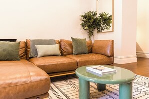 A brown leather sectional sofa with multiple cushions, a small round glass coffee table, and a patterned rug on a wooden floor.