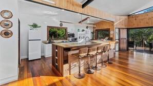 A modern kitchen with a central island, bar stools, and a view of the outdoors. - Large Beach Retreat with Ocean Breeze and Style (Hastings Point)