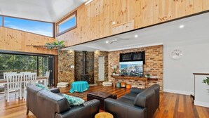 A modern living room with a stone fireplace, a television, and a dining area with wooden furniture. - Large Beach Retreat with Ocean Breeze and Style (Hastings Point)