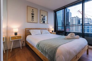 A bedroom with a wooden bed, white bedding, a green throw, bedside lamps, and a city view through large windows. - Skyline Escape I Chic City Stay + Free Parking (Melbourne)