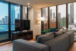 A modern living room with a large flat-screen TV, a sofa, a floor lamp, and a view of a cityscape through floor-to-ceiling windows. - Skyline Escape I Chic City Stay + Free Parking (Melbourne)