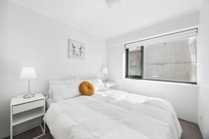A white bedroom with a bed, a nightstand, a lamp, a window, and a wall art piece.