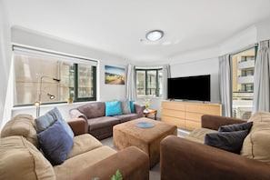 A modern living room with a sofa set, a coffee table, a flat-screen TV, and large windows with curtains.