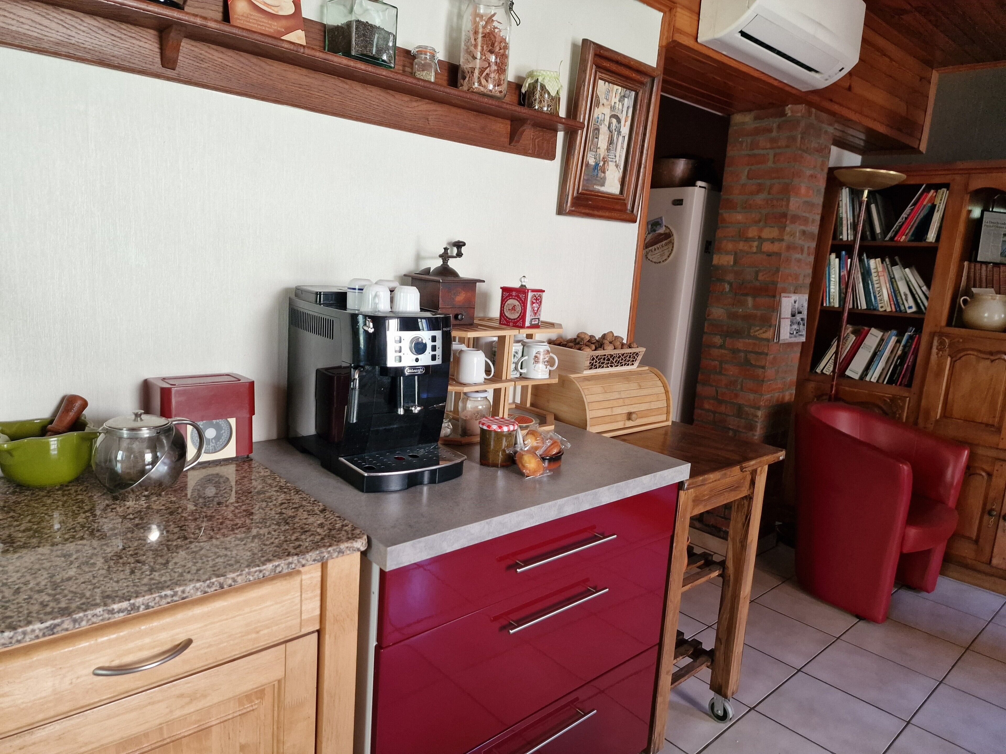 A kitchen with a coffee machine, a red cabinet, a red chair, and a bookshelf.