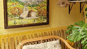Un cuadro enmarco que muestra un paisaje montañoso con una iglesia, una silla de mimbre y una planta en maceta.